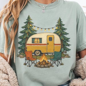 Peut inclure: Un t-shirt vert sauge avec une illustration représentant une caravane vintage jaune et blanche garée entre deux sapins. Une guirlande lumineuse est suspendue au-dessus, et un feu de camp brûle entre deux chaises pliantes orange et bleu sarcelle.