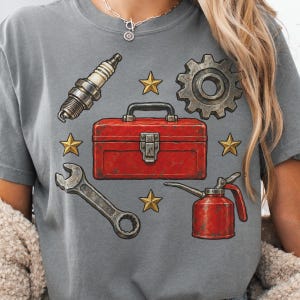 May include: A charcoal gray t-shirt featuring a graphic print of a red toolbox in the center, surrounded by a spark plug, a metal gear, a wrench, an oil can, and four gold stars arranged around the central toolbox illustration.