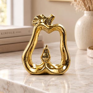 May include: A gold-colored ceramic sculpture shaped like an open-frame pear with a smooth, reflective surface. The decorative object features a hollow center and a small, solid pear-shaped accent positioned at the base within the frame.