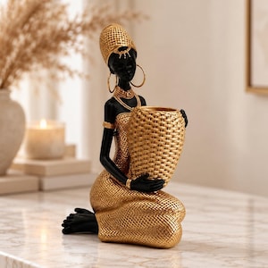 May include: A decorative figurine of a kneeling person with black skin, wearing a textured gold headpiece, large hoop earrings, and a matching gold dress. The figure holds a large, woven-style gold vessel in both hands.