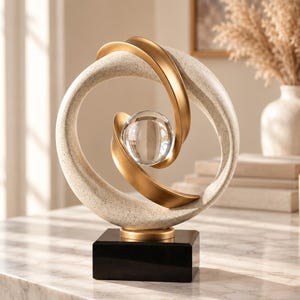 May include: A decorative sculpture featuring a textured beige circular frame with smooth gold accents, centered around a clear glass sphere. The entire piece is mounted on a solid black rectangular base, creating a balanced and structured composition.