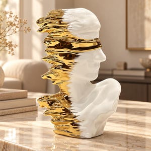May include: A white ceramic bust of a human head and shoulders featuring a blindfold. The left side of the sculpture transitions into a textured, metallic gold finish that appears to be melting or dripping down the side of the smooth white surface.