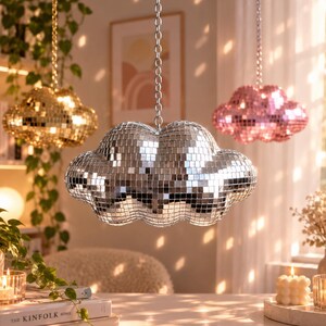 May include: Three cloud-shaped hanging decorations covered in small reflective mirror tiles. One cloud is gold, one is silver, and one is pink. Each cloud is suspended from the ceiling by a silver-colored metal chain, creating a sparkling effect.
