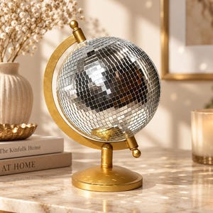 Puede incluir: Una bola de discoteca esférica montada sobre un soporte de metal dorado. La esfera está cubierta de pequeñas piezas de espejo plateadas y reflectantes. El soporte cuenta con una base circular, un brazo curvo y un eje central giratorio.