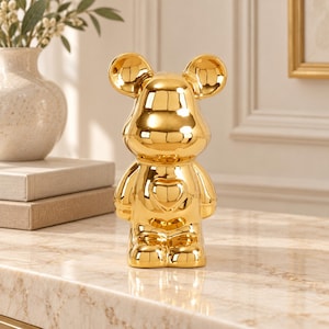 Peut inclure: Une figurine en forme d'ours de couleur or brillant avec une surface lisse et réfléchissante. L'ours présente des oreilles arrondies, une forme de tête distincte et un détail en forme de cœur centré sur sa poitrine, en position debout.