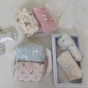 May include: A collection of six rectangular zippered pouches in various colors and patterns, including cream with teddy bears, solid pink corduroy, light blue heart-quilted fabric, and white with blue polka dots, each featuring a small decorative bow.