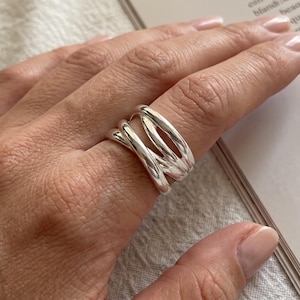 Puede incluir: Un anillo de plata pulida con un diseño de múltiples bandas que presenta tres hilos redondeados, lisos e interconectados que se superponen y se cruzan entre sí para crear una estructura ancha y abierta que rodea el dedo con un acabado reflectante.