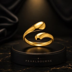 Puede incluir: Un anillo abierto de tono dorado con un acabado liso y pulido, que presenta dos extremos redondeados en forma de gota que no se tocan. El anillo se exhibe sobre una base circular negra con el texto PEARLBOURNE impreso en color blanco.