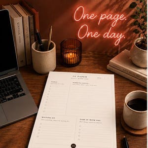 以下が含まれることがあります： IA PAPERと書かれた黒い文字の白いデイリーワークパッド。タスク、メモ、保留中、持ち出し用のセクションがあります。パッドは木製のデスクの上にあり、ノートパソコン、ペン立て、キャンドル、コーヒーカップが隣に置かれています。