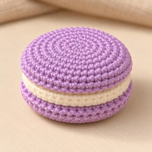 May include: A round, crocheted macaron-shaped object featuring two lavender-colored circular pieces with a thick, cream-colored center layer. The texture is created by tight, uniform yarn stitches, forming a layered, sandwich-like structure.