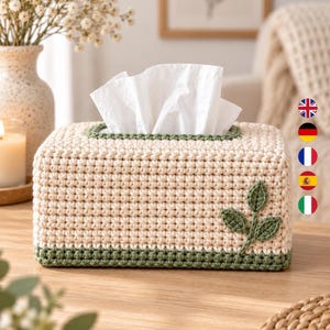 Puede incluir: Una funda rectangular para caja de pañuelos hecha de hilo de ganchillo color crema con un borde verde oscuro en la base y en la abertura superior. Una pequeña aplicación de ganchillo de tres hojas verdes está unida al frente.