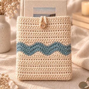 May include: A cream-coloured crochet sleeve for a book or tablet, featuring a horizontal blue wavy pattern across the centre. The sleeve is secured with a single light brown wooden button and a matching crochet loop closure at the top centre.