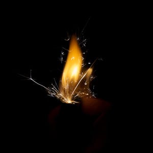 以下が含まれることがあります： 真っ黒な背景に、明るいオレンジ色の炎を放つ金属製のライター。炎の根元からは小さな白い火花が放射状に飛び散っており、コントラストの強い視覚効果を生み出しています。ライターの本体は暗闇の中に浮かび上がっています。