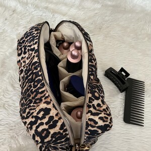 May include: A quilted leopard print travel case with a zipper closure, containing multiple hair styling attachments with metallic rose gold tops and a blue brush. A black plastic hair claw clip and a wide-tooth hair comb lie beside the case.