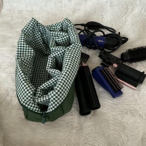May include: A dark green storage bag with a white and green gingham interior lining, accompanied by a blue Dyson hair styling tool, a black round brush, a black comb, and several black and blue hair styling attachments with metallic accents.