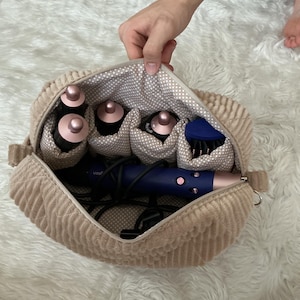 Könnte beinhalten: Eine beigefarbene Aufbewahrungstasche aus Kord mit Reißverschluss und einem beigeweißen, gepunkteten Innenfutter. Die Tasche enthält ein blaues Haarstyling-Gerät mit roségoldenen Akzenten sowie mehrere zylindrische Aufsätze in Einzeltaschen.