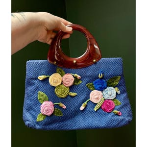 Op de afbeelding: Een blauwe gevlochten handtas met een donkerbruin gemarmerd handvat van hars en goudkleurige details. De voorzijde van de tas is versierd met diverse stoffen bloemapplicaties in roze, gele, groene en blauwe tinten, geaccentueerd met groene bladeren.