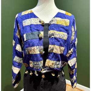 May include: A cropped jacket featuring a vibrant blue base with a patchwork design of rectangular fabric panels in various beige, brown, and patterned prints. The front closure consists of four toggle-style buttons with loops.