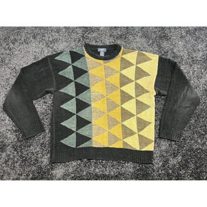 May include: A long-sleeve knit sweater featuring a geometric triangle pattern in shades of yellow, tan, sage green, and charcoal gray. The garment has a crew neckline, ribbed cuffs, and a ribbed hem, with solid charcoal gray sleeves and back.