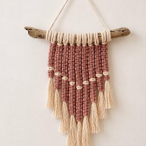 Puede incluir: Un tapiz de macramé con cordón de algodón en color rosa empolvado y crema anudado en un patrón escalonado. El tejido cuelga de una pieza de madera flotante natural con un lazo de cordón crema para colgar, acabado con borlas de color crema.