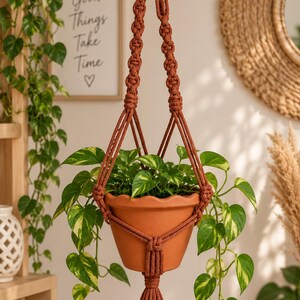 Op de afbeelding: Een terracotta-kleurige macramé plantenhanger met ingewikkelde geknoopte koordpatronen, een houten ring aan de bovenkant en een decoratieve kwast aan de onderkant, met een terracotta pot met geschulpte rand en een hangende groen-gele pothosplant.