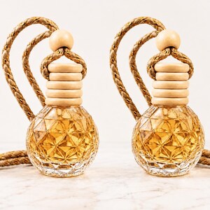 May include: Two identical glass car air freshener bottles with a geometric diamond-cut pattern filled with amber-colored liquid. Each bottle features a light-colored wooden cap and a thick, braided tan rope loop for hanging in a vehicle.