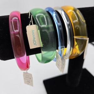 May include: A set of five translucent bangle bracelets in pink, green, blue, silver, and yellow. Each bracelet features a small, rectangular, silver-toned metallic accent embedded on the surface. A JCPenney tag is attached to the green bracelet.