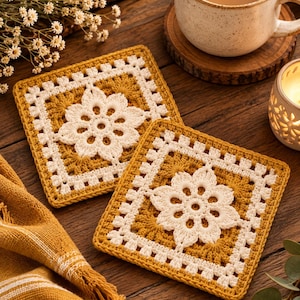 May include: Two square crochet coasters featuring a central white floral motif surrounded by mustard yellow yarn. The square coasters have a white border with a repeating open-stitch pattern, creating a textured, geometric design for table surfaces.