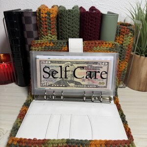 May include: A crochet binder cover featuring a variegated autumn-toned yarn in shades of orange, brown, and green. The binder includes a white interior with card slots and a clear plastic insert displaying a fifty-dollar bill labeled Self Care.