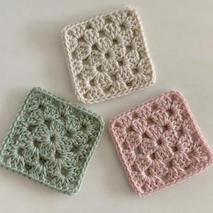 May include: Three square crochet granny squares arranged in a cluster. One square is cream, one is sage green, and one is light pink. Each piece features a central floral-inspired pattern created with yarn, showcasing a classic openwork stitch design.