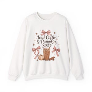 May include: A white crewneck sweatshirt featuring a graphic print of a glass of iced coffee, a pumpkin, and a cinnamon stick. The design includes pink bows and small stars, with the text "Iced Coffee & Pumpkin Spice" in dark brown script.