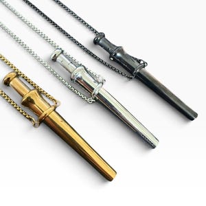 May include: Three pendant necklaces featuring a miniature sword-like design with a sheath. The pendants are finished in gold, silver, and dark gunmetal tones, each suspended from a matching metallic chain that attaches to the hilt and sheath.