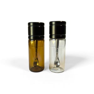 May include: Two cylindrical glass vials with black screw-top lids, each containing a small silver-toned metal spoon attached to the underside of the cap. One vial is made of amber-colored glass, while the other is made of clear, transparent glass.