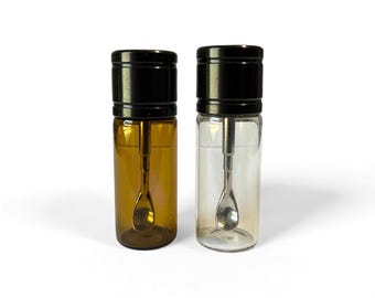 May include: Two cylindrical glass vials with black screw-top lids, each containing a small silver-toned metal spoon attached to the underside of the cap. One vial is made of amber-colored glass, while the other is made of clear, transparent glass.