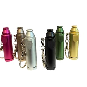 May include: A collection of six cylindrical metal pill case keychains in various colors including magenta, bronze, silver, black, dark green, and gold. Each keychain features a screw-top lid, a small shield-shaped emblem, and a metal split ring.