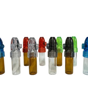Puede incluir: Un conjunto de diez pequeños dispensadores cilíndricos de rapé dispuestos en grupo. Cada dispensador cuenta con una base de frasco de vidrio transparente o ámbar rematada con una tapa de plástico de colores, incluyendo variantes en azul, transparente, rojo, negro y verde.
