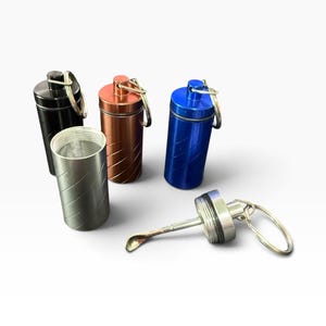May include: A collection of four cylindrical metal containers with screw-top lids and attached key rings. The containers come in black, copper, blue, and silver, each featuring a diagonal grooved pattern. One silver lid is detached, revealing a small spoon.