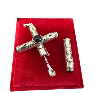 May include: A silver cross pendant featuring a central black stone, intricate circular metal patterns, and a detachable cap that reveals a small, thin metal spoon attached to the interior. The cross has a bail at the top for hanging.