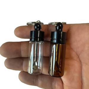 以下が含まれることがあります： 黒いスクリューキャップと金属製のキーリングが付いた、2つの小さな円筒形のガラス瓶。1つは透明なガラス、もう1つは琥珀色のガラスです。それぞれの瓶の中には、小さな銀色の金属製スプーンが入っています。