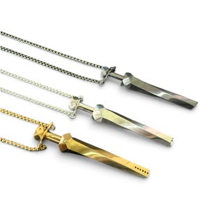 May include: Three sword-shaped pendants on metal chains. The top pendant is dark grey, the middle is silver-toned, and the bottom is gold-toned. Each pendant features a faceted, twisted blade design with a crossguard and a thin, linked chain.