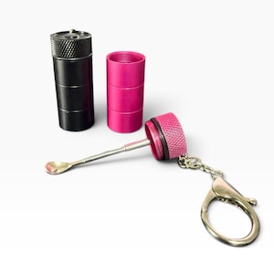 Puede incluir: Un recipiente cilíndrico negro, un recipiente cilíndrico rosa y una tapa rosa con una cuchara de metal adjunta y un anillo de llavero plateado. La tapa presenta un patrón de agarre moleteado texturizado y la cuchara está conectada por una varilla metálica.