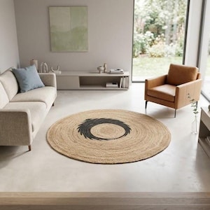 May include: A round, woven jute rug featuring a natural tan color with a dark charcoal black circular pattern in the center. The rug is positioned on a light-colored floor in a room containing a beige sofa, a tan leather armchair, and a console.