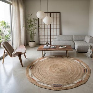 May include: A round, woven jute rug featuring a natural tan color with decorative geometric border accents in white, orange, and blue. The rug is textured with a concentric circular pattern and sits on a smooth, light-colored concrete floor.