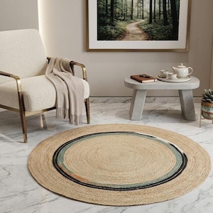 May include: A round, woven jute rug featuring concentric circles in natural tan, muted teal, and black. The rug is placed on a white marble floor in front of an armchair with a beige throw blanket, a concrete side table, and wall art.