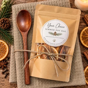 May include: A brown paper pouch labeled Your Choice SIMMER POT BAG with a clear window showing dried orange slices and cinnamon sticks, tied with twine to a dark wooden spoon. The label features a pinecone graphic and text including Mamas Modern Manor.