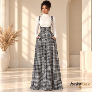 May include: A white long-sleeved turtleneck top paired with a high-waisted, floor-length black and white gingham skirt featuring suspender straps and a central vertical row of buttons. The skirt includes side pockets and a full silhouette.