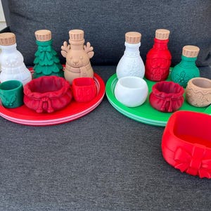 May include: A collection of holiday-themed silicone items including two round trays, various small bottles with stoppers, miniature bowls, and a red square container with a bow design. The items feature festive shapes like snowmen, trees, reindeer, and Santa.