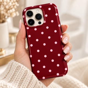 Könnte beinhalten: Eine dunkelrote Smartphone-Hülle mit einem sich wiederholenden Muster aus kleinen, hellrosa Punkten. Die Hülle hat eine Aussparung für ein Drei-Linsen-Kamerasystem auf der Rückseite, eine glänzende Oberfläche und abgerundete Ecken.