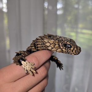 Peut inclure: Une figurine de lézard articulée imprimée en 3D avec un motif d'écailles bronze métallique et noir. La créature possède un corps segmenté texturé avec des écailles détaillées, une tête pointue aux yeux sombres et des pattes griffues.