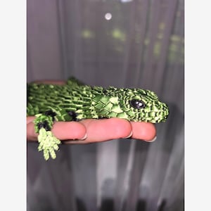 Peut inclure: Une figurine de lézard vert conçue avec une surface texturée rappelant des écailles. Le lézard présente une tête pointue, des yeux ovales sombres et des pattes griffues détaillées. Le corps est composé de segments verts superposés avec des accents noirs.